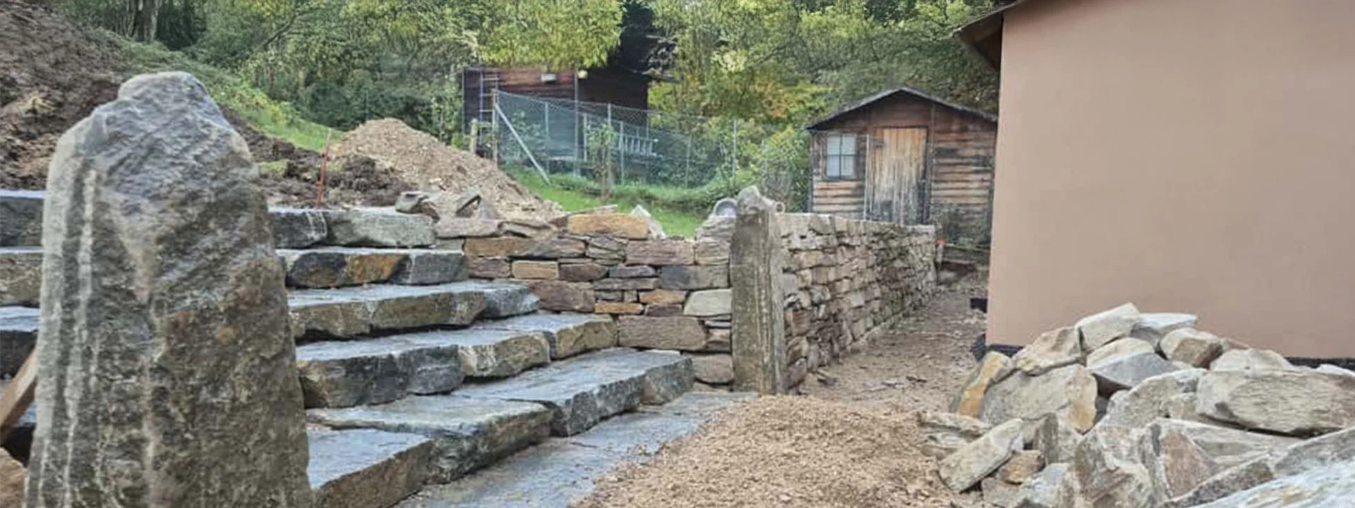 Natursteintreppe und Trockenmauer im Bau, umgeben von Erdarbeiten und Gartenfläche.